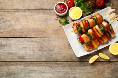 Photo of Delicious chicken shish kebabs with vegetables and ketchup on wooden table, flat lay. Space for text