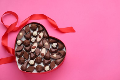 Heart shaped box with delicious chocolate candies and ribbon on pink background, flat lay. Space for text