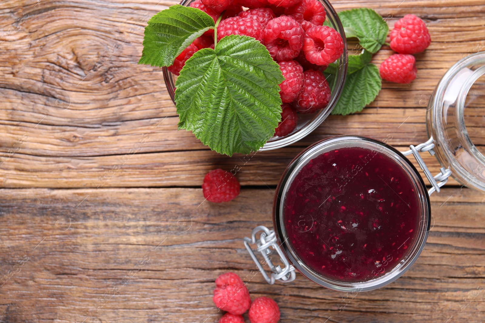 Photo of Jar of delicious raspberry jam, fresh berries and green leaves on wooden table, flat lay. Space for text