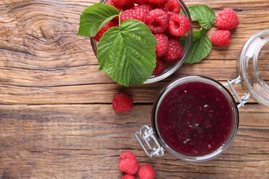 Jar of delicious raspberry jam, fresh berries and green leaves on wooden table, flat lay. Space for text