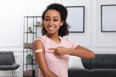 Photo of Happy young woman pointing at adhesive bandage after vaccination indoors