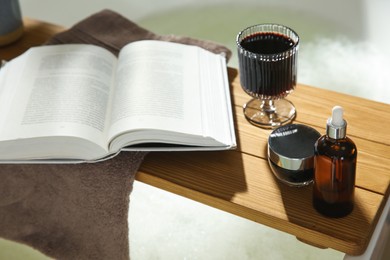 Wooden bath tray with open book, glass of wine and cosmetic products on tub indoors