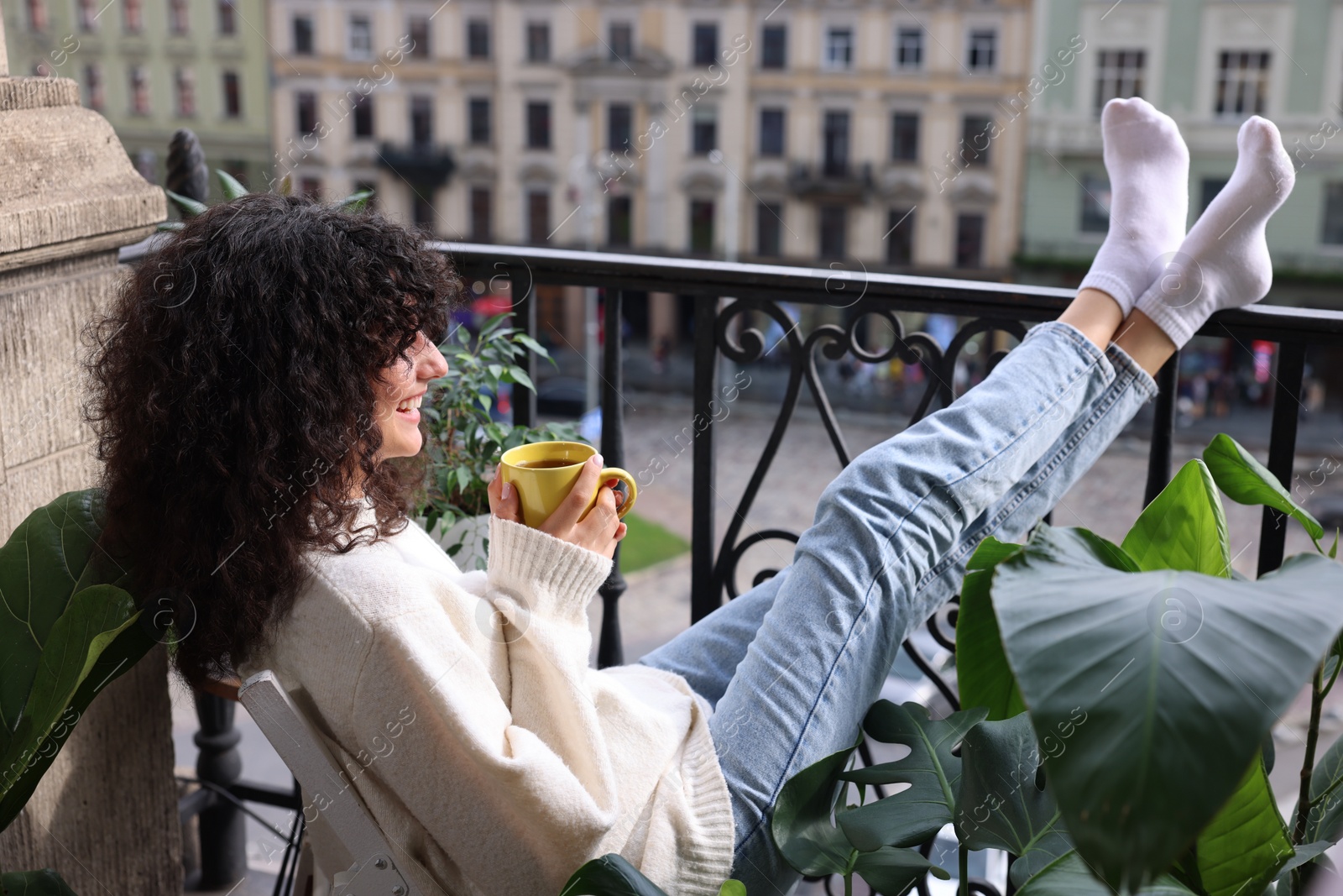 Photo of Young woman with cup of tea relaxing in chair surrounded by green houseplants on balcony