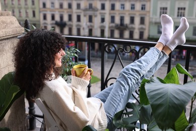 Photo of Young woman with cup of tea relaxing in chair surrounded by green houseplants on balcony
