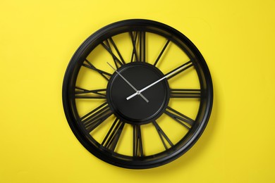 Photo of Modern black clock with Roman numerals on yellow background, top view
