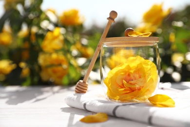 Glass jar with yellow rose and honey dipper on white wooden table in blooming garden, space for text