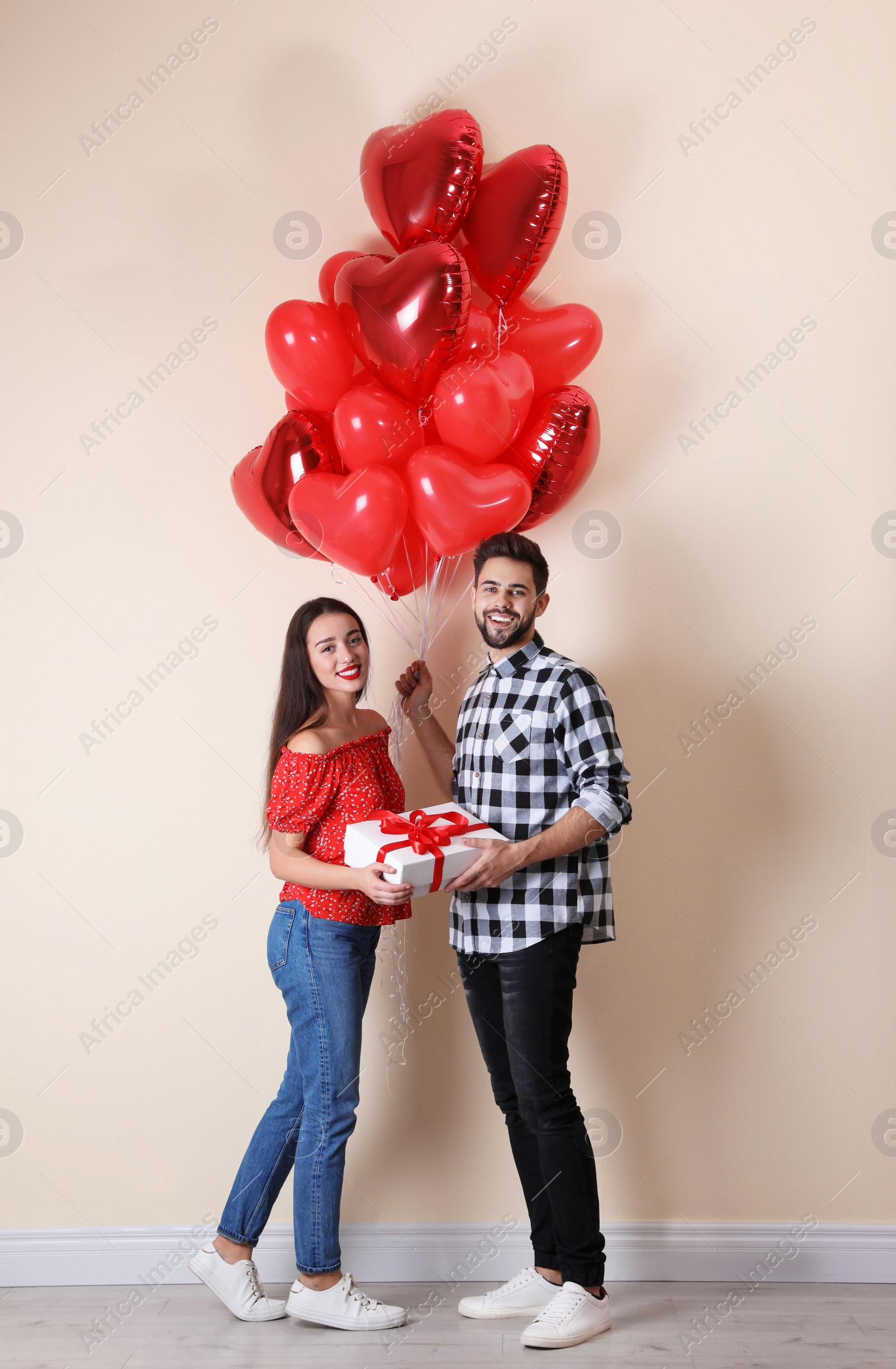 Photo of Happy young couple with gift box and heart shaped balloons near beige wall. Valentine's day celebration