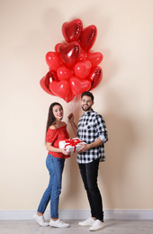 Photo of Happy young couple with gift box and heart shaped balloons near beige wall. Valentine's day celebration
