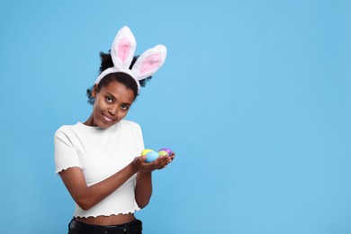 Photo of Happy African American woman in bunny ears headband holding Easter eggs on light blue background. Space for text