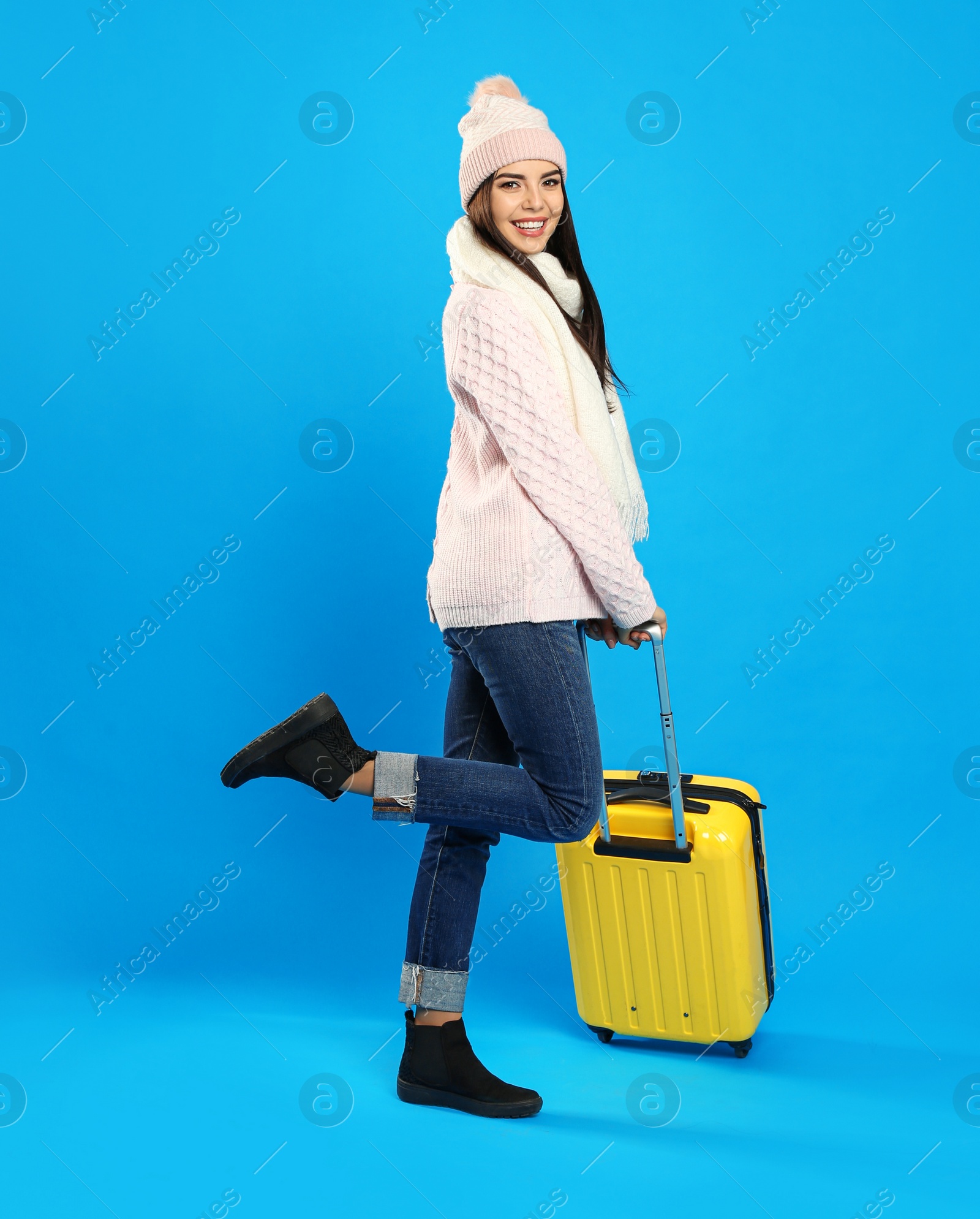 Photo of Young woman in warm clothes with suitcase on blue background. Winter vacation