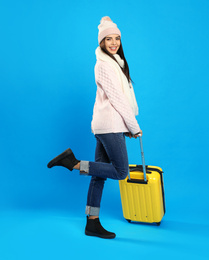 Young woman in warm clothes with suitcase on blue background. Winter vacation