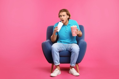 Emotional man with popcorn and beverage sitting in armchair during cinema show on color background