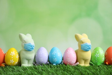 Image of COVID-19 pandemic. Easter bunnies in protective masks and painted eggs on green grass