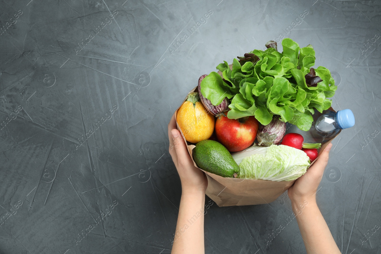 Photo of Woman holding paper package full of fresh vegetables and fruits on dark background, top view. Space for text