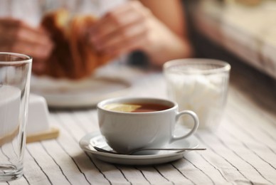 Photo of Woman eating croissant at table, focus on cuparomatic tea with lemon