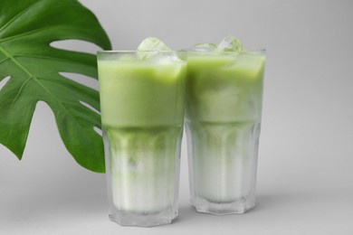 Glasses of tasty iced matcha latte and leaf on light grey background