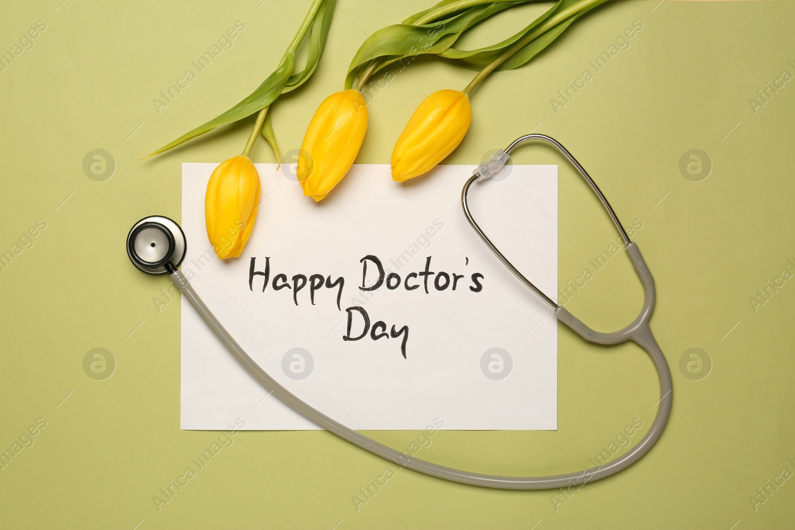 Photo of Card with phrase Happy Doctor's Day, stethoscope and yellow tulips on light green background, flat lay