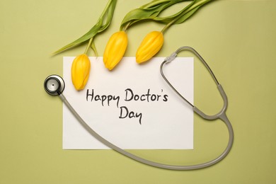 Card with phrase Happy Doctor's Day, stethoscope and yellow tulips on light green background, flat lay