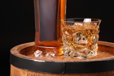 Photo of Whiskey in glass, bottle and ice cubes on wooden barrel against black background