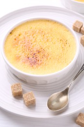 Photo of Delicious creme brulee in bowl, sugar cubes and spoon on white table