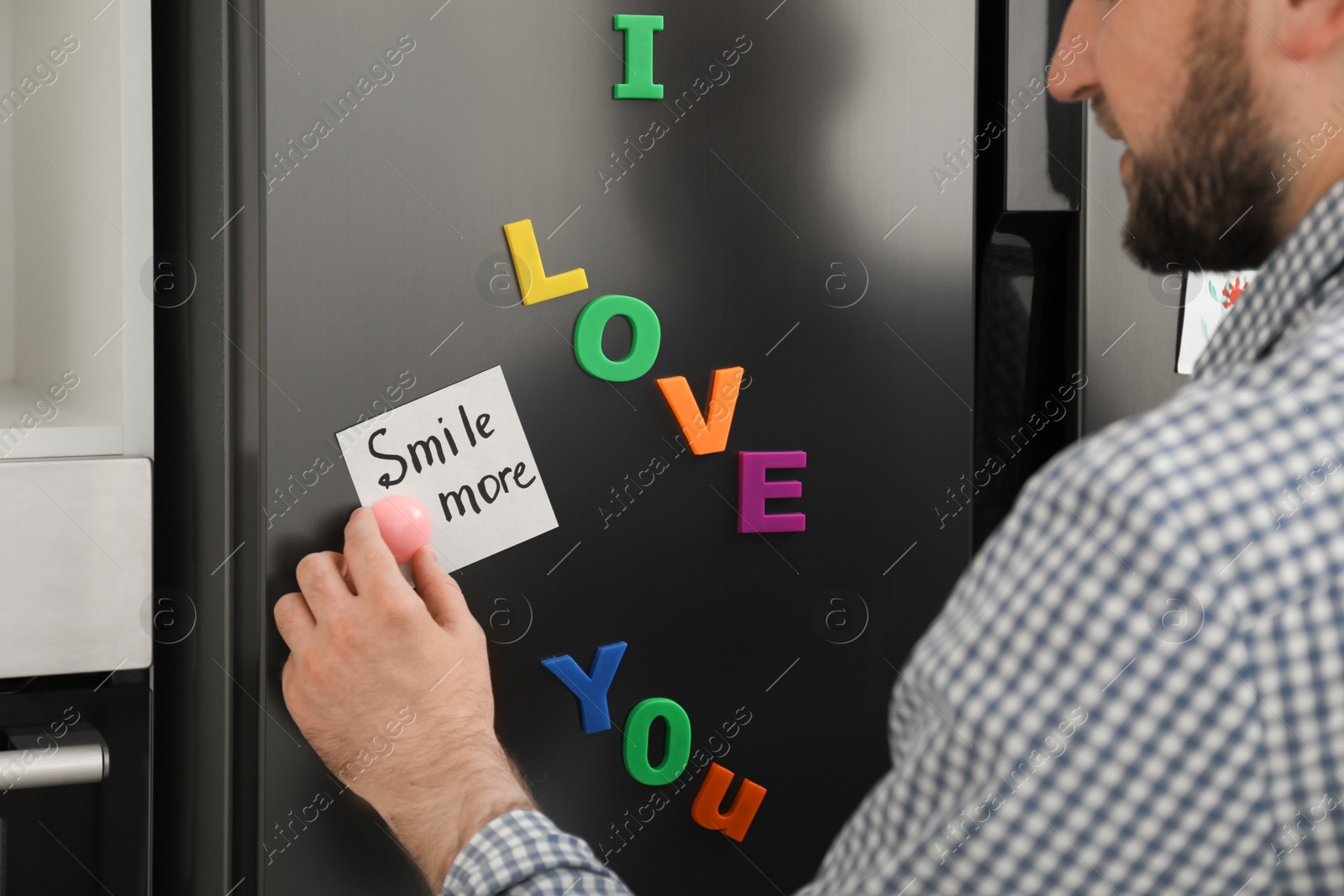 Photo of Man putting note with phrase SMILE MORE on refrigerator door, closeup