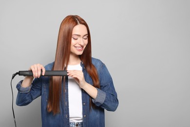 Beautiful woman using hair iron on light gray background, space for text