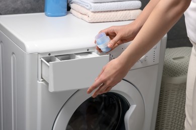 Photo of Woman pouring fabric softener from cap into washing machine, closeup