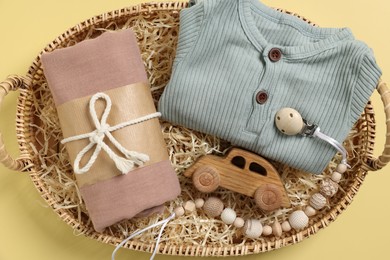 Different baby accessories and clothes in wicker basket on yellow background, top view