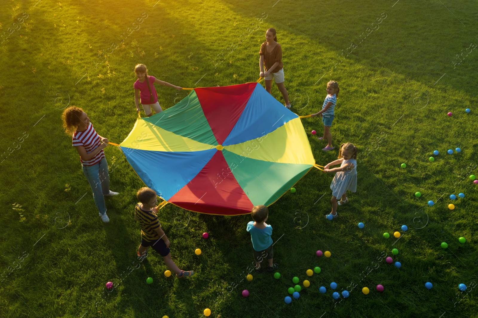 Image of Group of children and teachers playing with rainbow playground parachute on green grass, above view. Summer camp activity