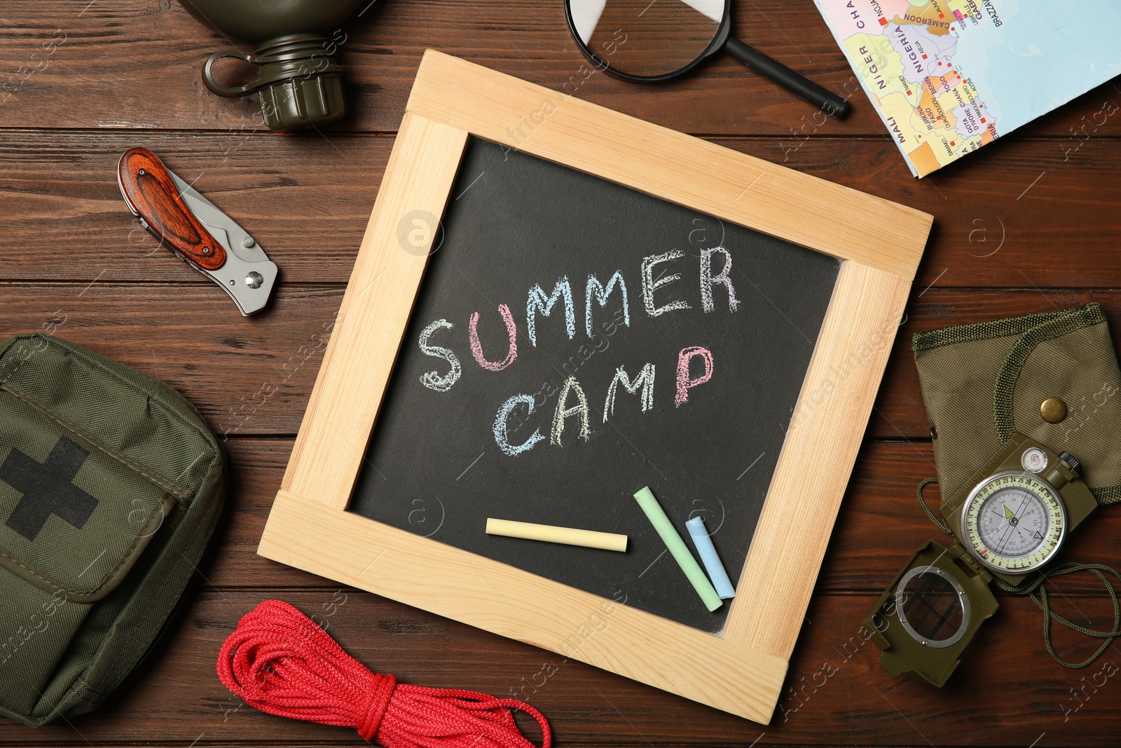 Photo of Chalkboard with text SUMMER CAMP and camping equipment on wooden background, flat lay