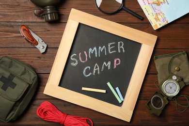 Chalkboard with text SUMMER CAMP and camping equipment on wooden background, flat lay