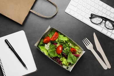 Photo of Container of tasty food, keyboard, cutlery and notebook on grey table, flat lay. Business lunch