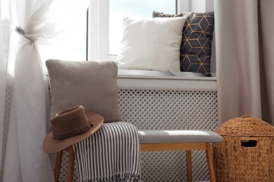 Photo of Striped plaid, hat and cushion on bench near window sill with pillows in room. Cozy place to relax