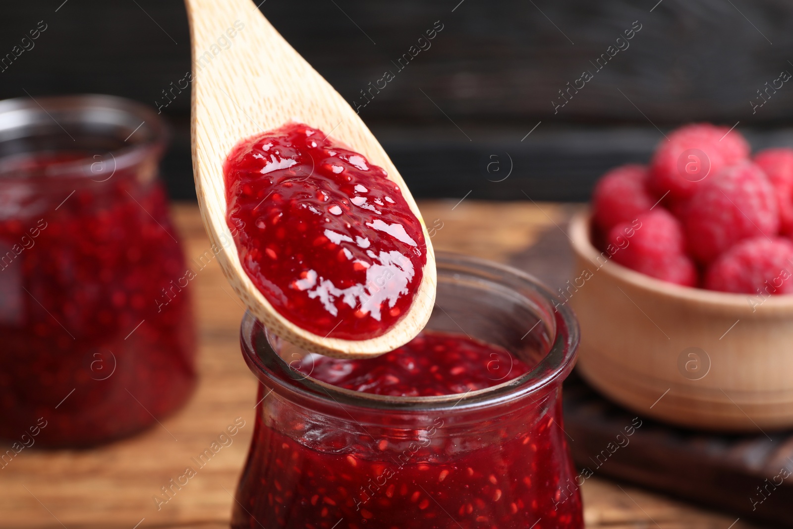 Photo of Spoon with raspberry jam over glass jar on table, closeup