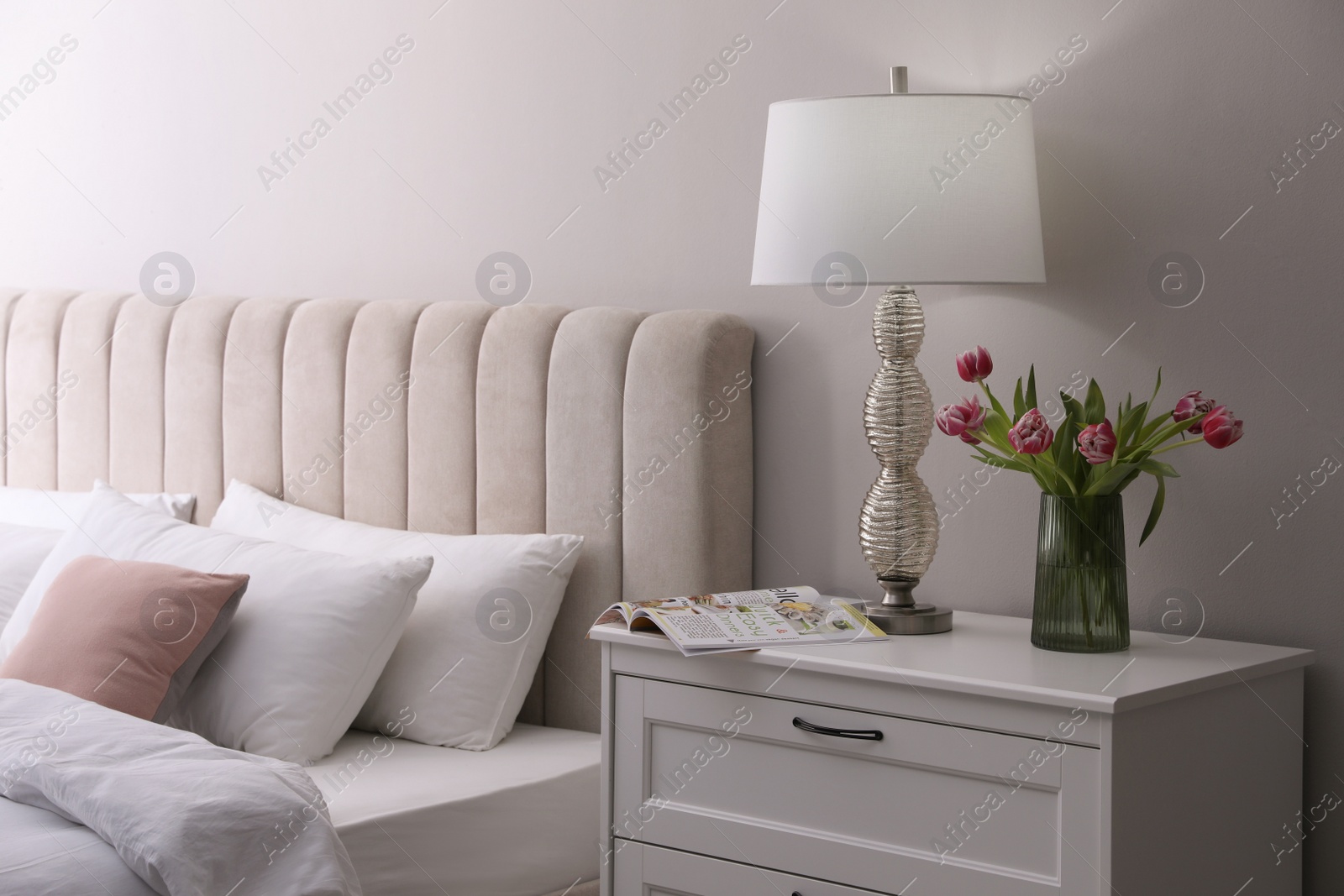 Photo of Stylish lamp, flowers and magazine on bedside table indoors. Bedroom interior elements