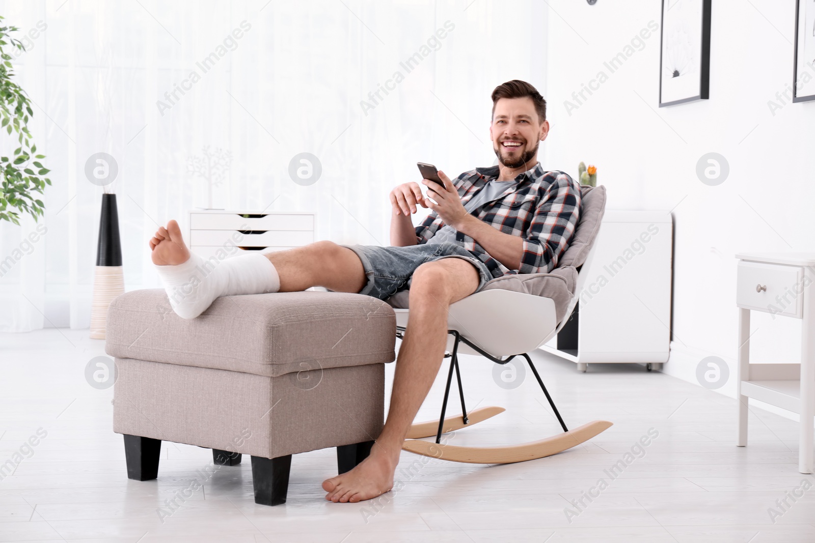 Photo of Man with broken leg in cast using mobile phone while sitting in armchair at home