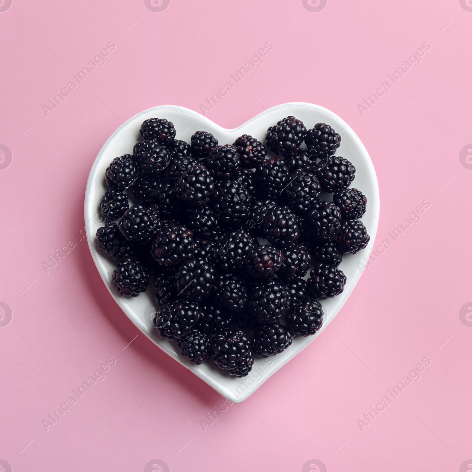 Photo of Heart shaped plate of ripe blackberries on pink background, top view