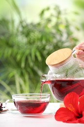 Photo of Woman pouring delicious hibiscus tea into cup at white wooden table outdoors, closeup