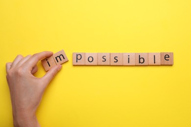 Photo of Motivation concept. Woman changing word from Impossible into Possible by removing wooden squares on yellow background, top view