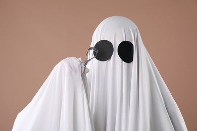 Fashionable ghost. Woman covered with white sheet using eyelash curler on dark beige background