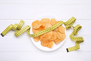 Plate with potato chips and measuring tape on white wooden table, flat lay. Weight loss concept