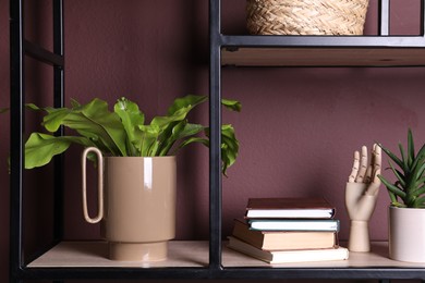 Stylish ceramic vase with green leaves, books and decor on shelf near brown wall