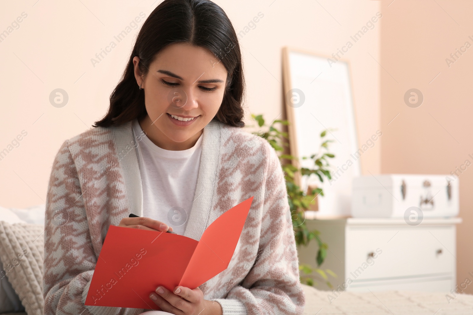 Photo of Young woman writing message in greeting card indoors, space for text