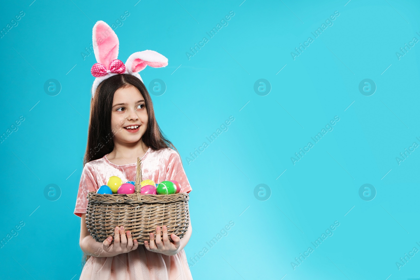Photo of Little girl in bunny ears headband holding basket with Easter eggs on color background, space for text