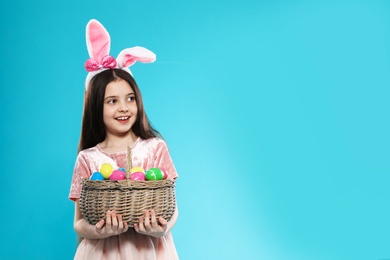 Photo of Little girl in bunny ears headband holding basket with Easter eggs on color background, space for text