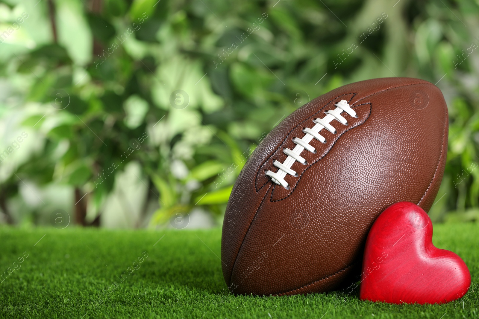 Photo of American football ball and red heart on green grass against blurred background. Space for text