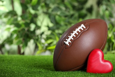 Photo of American football ball and red heart on green grass against blurred background. Space for text