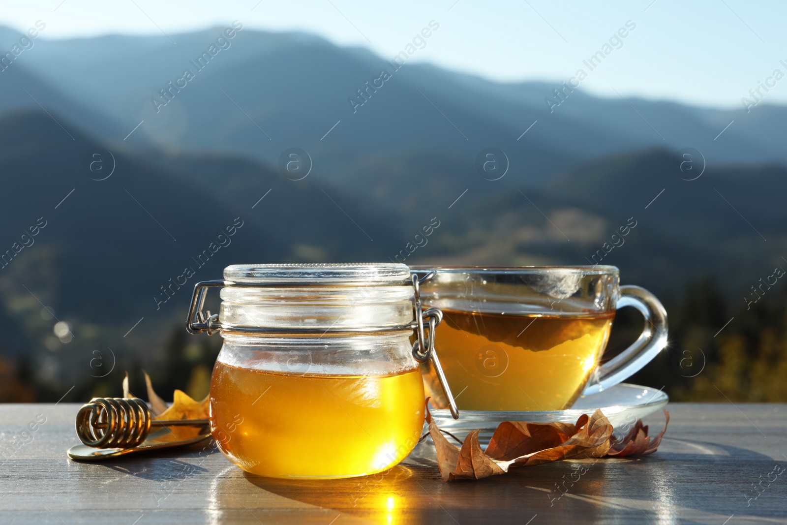 Photo of Fresh aromatic honey and cup of hot tea on grey wooden table against mountain landscape