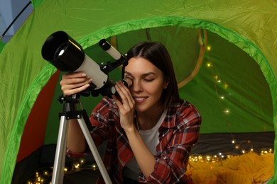 Photo of Young woman looking at stars through telescope while sitting in camping tent indoors
