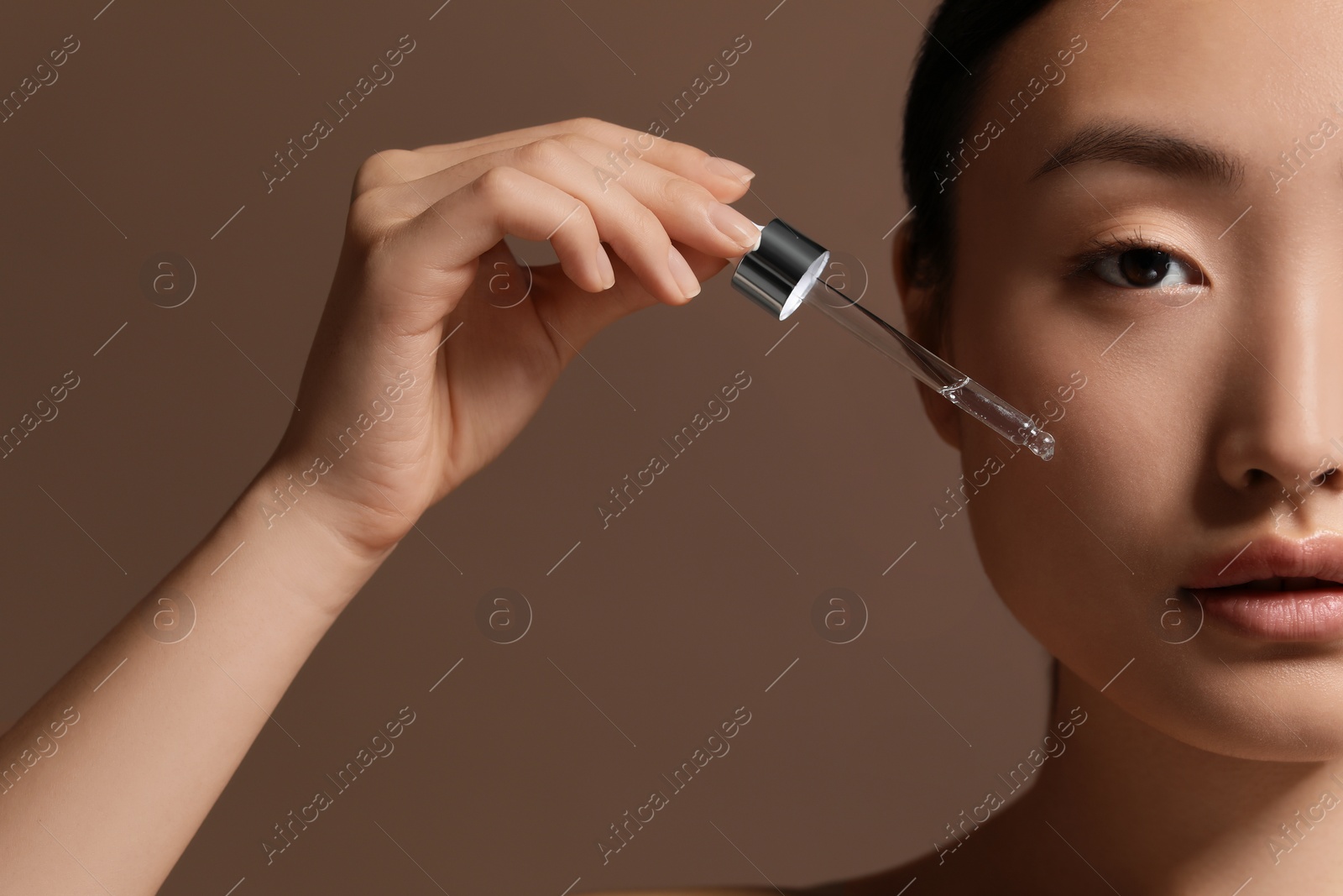 Photo of Beautiful young woman applying cosmetic serum onto her face on brown background, closeup